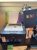 Full view of Brother HL-2230 printer on desk with multipurpose paper pack on top and box with supplies nearby.