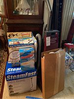 View of boxed items including Steadfast fabric steamer, DampRid moisture absorber, Dryer Max lint removal kit, and Hoover Legacy vacuum unit with hose, spare parts and vacuum bags visible in background along with metal laundry sign