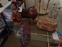 Lot containing wood carved masks, a pedestal jar, a corn husk doll, decorative box, metal butterfly lamp and plastic Hawaiian souvenir as seen on glass table.