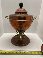Front view of antique copper and brass samovar showing rounded body, brass side handles, domed lid with black knob, and brass spigot with black handle. Measuring tape at base shows about 11-12 inches width.