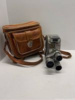 Side view of the Keystone 27 Capri 8mm camera beside its brown leather carrying case on a white background.