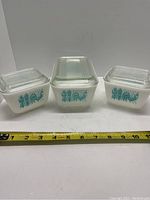 Three white opaque Pyrex glass containers with clear ribbed lids, all with blue Amish Butter roosters and wheat design, ruler for scale in front.