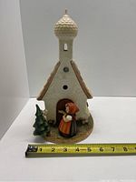 Front view of vintage Hummel figurine clock housing featuring a white building with a dome roof, girl figurine, and tree next to a measuring tape for size reference.