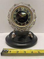 Front view of the vintage Majak table clock showing faceted clear glass case, brown dial with gold numbers, and black oval base.