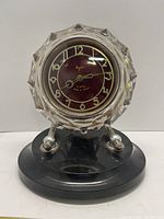Full front view of the Majak clock showing burgundy dial, faceted glass cover, metal stands, and black base.