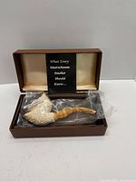 Open brown rectangular case with cream satin interior holding meerschaum pipe in plastic sleeve and informational pamphlet