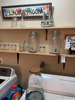 Glass bottles of various sizes arranged on wooden shelves above laundry machines, with decorative laundry signs on the wall.