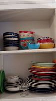 Shelf with stacked blue and white striped bowls, colorful floral and polka dot bowls, clear small glass dessert bowl, and stacked plates