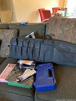 Black nylon carrying case with multiple pockets and two handles placed on a couch with various cleaning items around it including a blue plastic box and cleaning supplies.