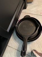 Two cast iron skillets stacked on the floor, top one showing the distinctive handle shape of the Pioneer Woman skillet