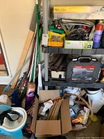 Photo shows shelving unit with Husky socket set case, yellow Buck Bros box, assorted cleaning and repair supplies, cardboard boxes with miscellaneous items and bulbs, mop bucket with wringer, and multiple cleaning tools leaning against the wall.