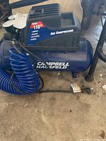 Front and side view of Campbell Hausfeld portable air compressor with coiled blue air hose on concrete floor.