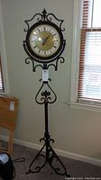 Full view of metal floor clock, showing its height, stand and clock face in a room corner near windows.