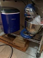 Blue Della ice maker, blue KitchenAid mixer, stoneware bar pan and Pampered Chef coating trays on metal shelving