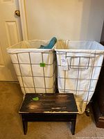 Front view of two white cloth laundry hampers with metal frames on wheels, alongside a small wooden stool with worn black paint finish.