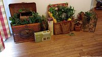 Longaberger trunk, picnic basket, faux flowers, faux fruit, wine rack, glass floor vase, planter with faux flowers, box of grape lights, and wine glasses arranged on wooden floor against a wall.