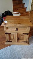 Front angled view of wooden nightstand showing drawer and cabinet doors with metal hardware in a living room setting with hardwood floors and carpet.