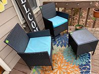 Front left view of the outdoor black wicker-style table and two matching armchairs with blue cushions, placed on a deck with a colorful patterned rug beneath.