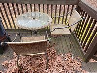 View showing round outdoor table with glass top and wicker trim, two wicker-seated chairs on wooden deck with fallen leaves.