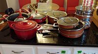 Multiple pots and pans, mostly red-colored with glass lids and stainless steel handles, on a black countertop.