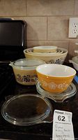 Pyrex mixing bowls and baking dishes in Homestead pattern with orange bowl and glass lids stacked on kitchen counter.