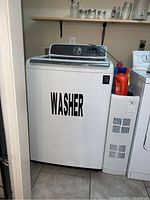 Wide view of white Samsung top loading washing machine in laundry room, showing front and control panel with 'WASHER' label on front