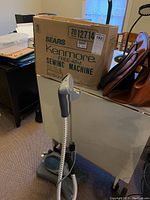 Photo showing the Kenmore 14 sewing machine original box, fabric steamer with hose and handle, and brown sewing machine case on a white table surface.