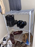 White metal wire shelving unit with three tiers placed in a corner with shoes on and around it.