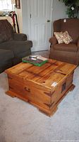 Front and side view of wood coffee table showing natural wood finish, metal corner brackets, and latch on the front. Table sits on carpeted floor with couches behind it.