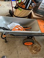Side and front view of the yard cart with gray basin and orange wheels, with some dirt inside the basin and orange metal handle pieces resting inside.