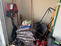 View of multiple yard tools leaning against garage wall, including bags of mulch (mulch not included), metal hand tools, wheelbarrow partially visible
