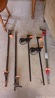 Five bar clamps laid on carpet; two long rusty metal clamps, one rusty clamp missing part of handle, two black and orange plastic handle clamps