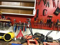 Hand tools hanging on red pegboard wall including clamps, pliers, assorted screwdrivers, clamps, and other hand tools along with metal yardstick and level on shelves.