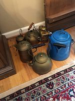 Photo showing a cluster of copper kettles, watering cans, and a blue porcelain coffee pot on a hardwood floor beside a rug.