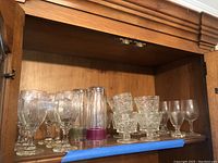 Glassware set on wooden shelf including stemware wine glasses, cut glass bowls, and tall juice glasses.