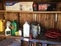 Overall photo of shelf showing Havahart live animal trap box, Flo Tool drain container, gas and oil containers, red jumper cables coiled, and other garden and automotive supplies.