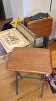 Tray tables standing and stacked: Four wooden trays on a wooden stand, front metal tray table with natural wood finish and folding legs.