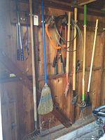 Full view of all yard tools hanging on the wooden wall inside a shed. Showing broom, pry bar, rakes, clippers, shovel, and saw.