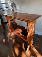 Wooden library style side table with cut-out side supports and a lower shelf for storing books, lit by natural sunlight.