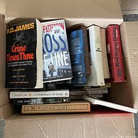 Top view of open box showing a collection of paperback and hardcover books including Crime Times Three by P.D. James, Cross Line by Patricia Cornwell, and others.