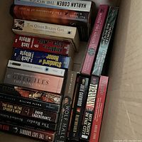 Box containing about 20 mixed books showing various titles such as 'The Woods', 'The Other Boleyn Girl', '1984', and 'Wolf Hall'.