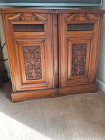 Front view of wooden cabinet showing carved floral designs on two doors and decorative fluted panel above each door panel.