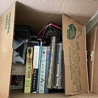 Overhead view of box with a variety of books including titles from authors like James Patterson and Laurie R. King, some modern paperbacks visible.