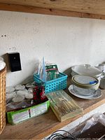 Glass jars with white lids, red and black container, blue basket with utensils, packaged cutlery set, and plates on a wooden shelf.