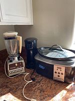 Photo of three kitchen appliances on counter with sunlight: Osterizer blender, black coffee maker, and Crockpot slow cooker with glass lid