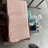 Photo showing a pink plastic dish drying rack wrapped in plastic, a white soap dispenser with pump, packages of disposable plastic forks and spoons, and various small unopened items including packaged tissues and cleaning gloves.