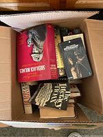Photo showing a cardboard box filled with multiple books, some titles visible such as Sherlock Holmes, mythology book, and others.