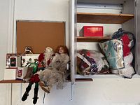 View of various Christmas decorations including dolls, boxed figurines, stocking, and bags with decorations on shelves and hanging on corkboard.