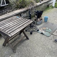 Outdoor wooden folding patio table with slatted top placed on concrete. Surrounding items include several metal pot holders, some ceramic pots, and a decorative rubber doormat with floral design.