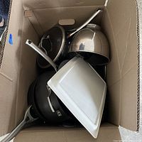 Box containing various metal pots and pans along with a white rectangular baking tray.